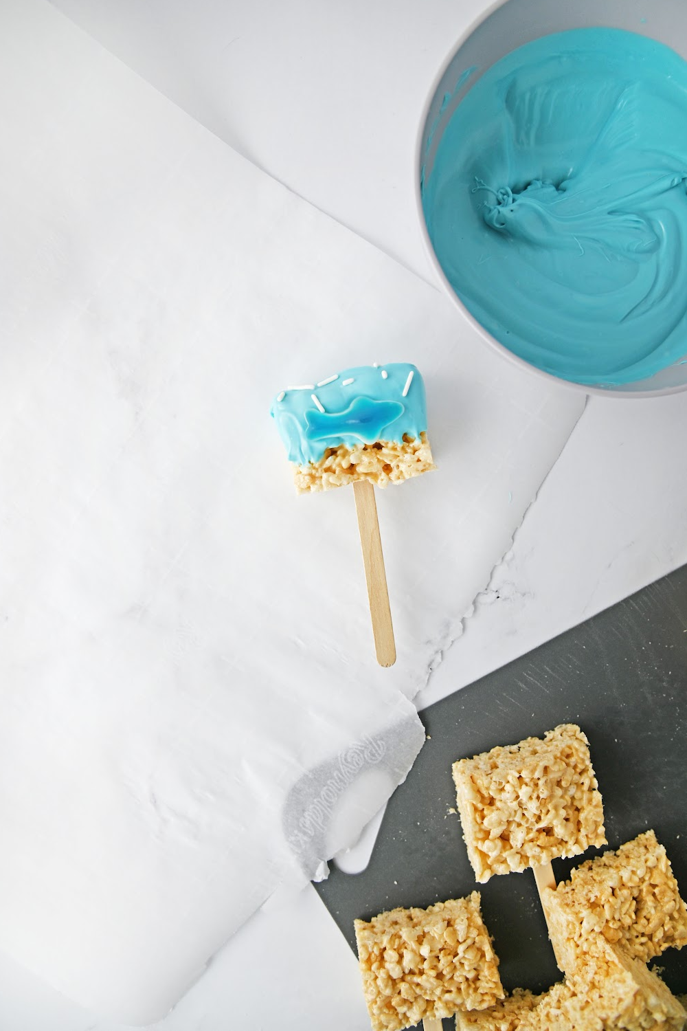 1 shark Rice Krispie treat pops made with a gummy shark laying on parchment paper with undecorated rice Krispie treats.