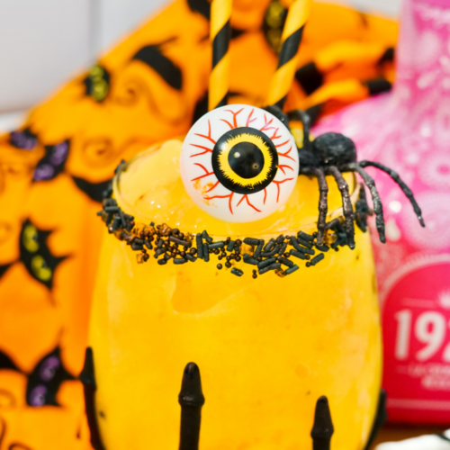 Orange mango margarita with black sprinkles rimming the glass. This Halloween margarita is garnished with a spooky eyeball and a black spider!