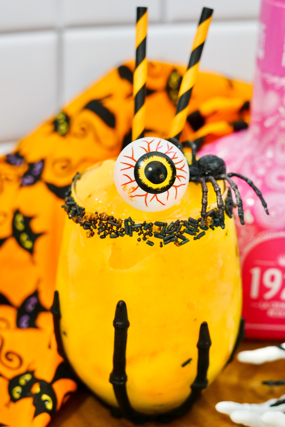 Orange mango margarita with black sprinkles rimming the glass. This Halloween margarita is garnished with a spooky eyeball and a black spider!