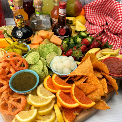 A margarita charcuterie board. A variety alcohol and mixers is displayed on one corner of the board. An assortment of zesty chips, peppers and fruits fill the rest of this unique charcuterie platter.