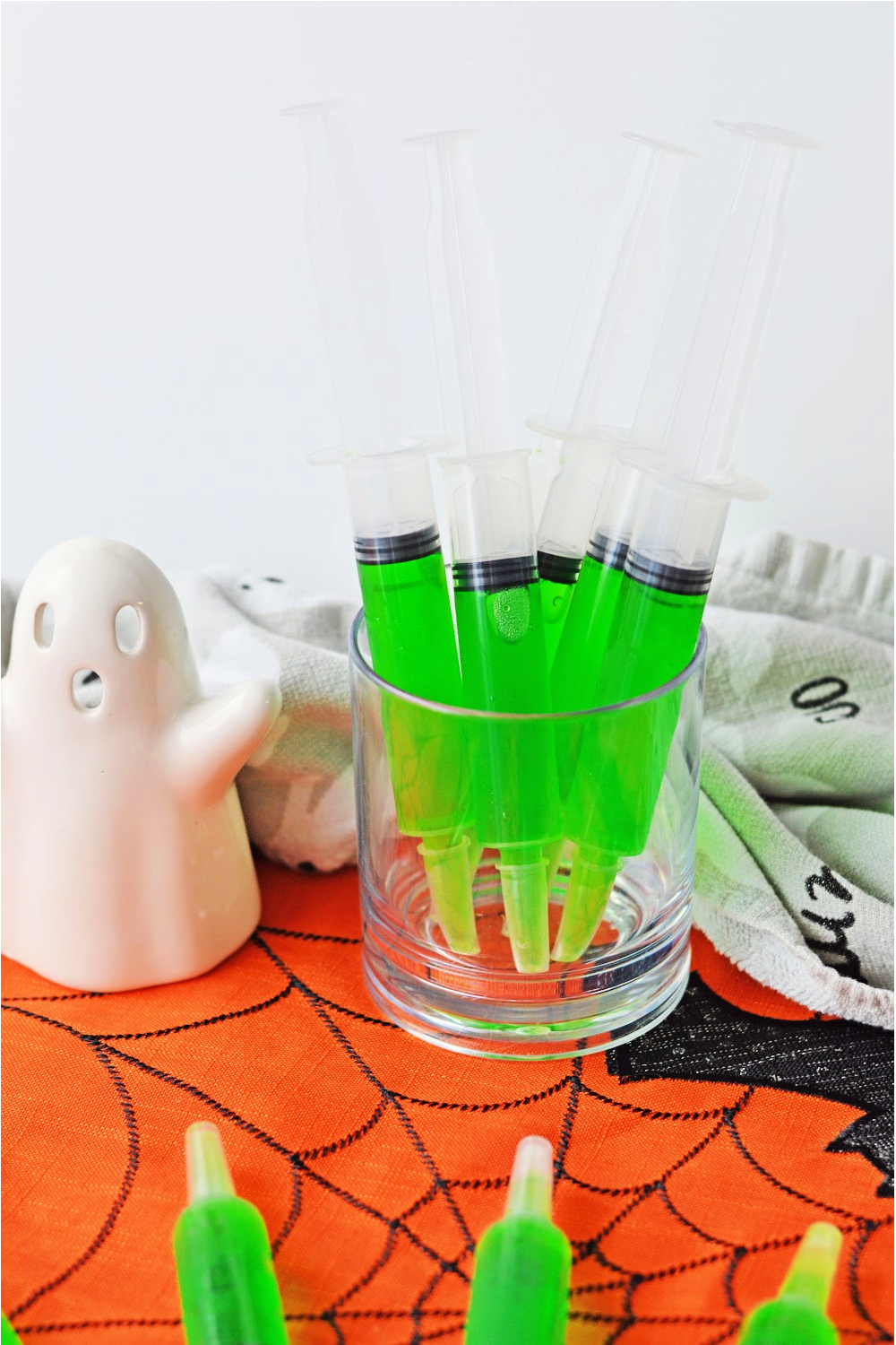 a glass container is holding the green syringe jello shots sitting on an orange Halloween placemat.