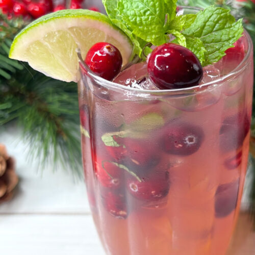 A pinterest image with a holiday mojito garnished with cranberries, mint and a wedge of lime. Winter greens are seen in the background.