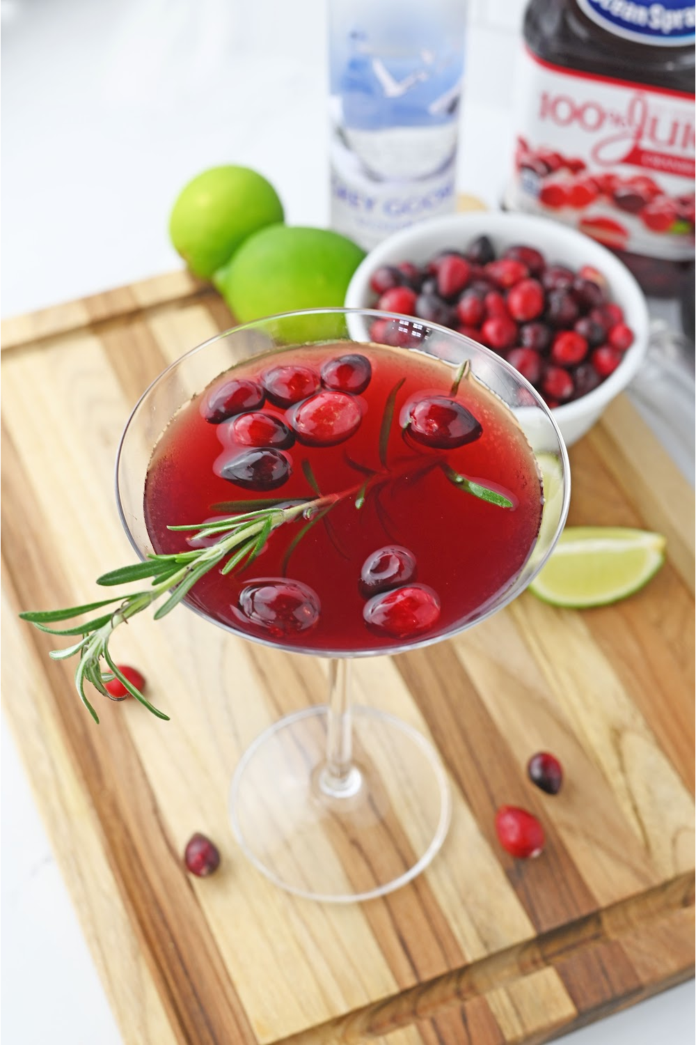 delightful Christmas Martini cocktail recipe garnished with fresh cranberries and a sprig of rosemary. Glass is sitting on a cutting board with slice limes and other ingredients.