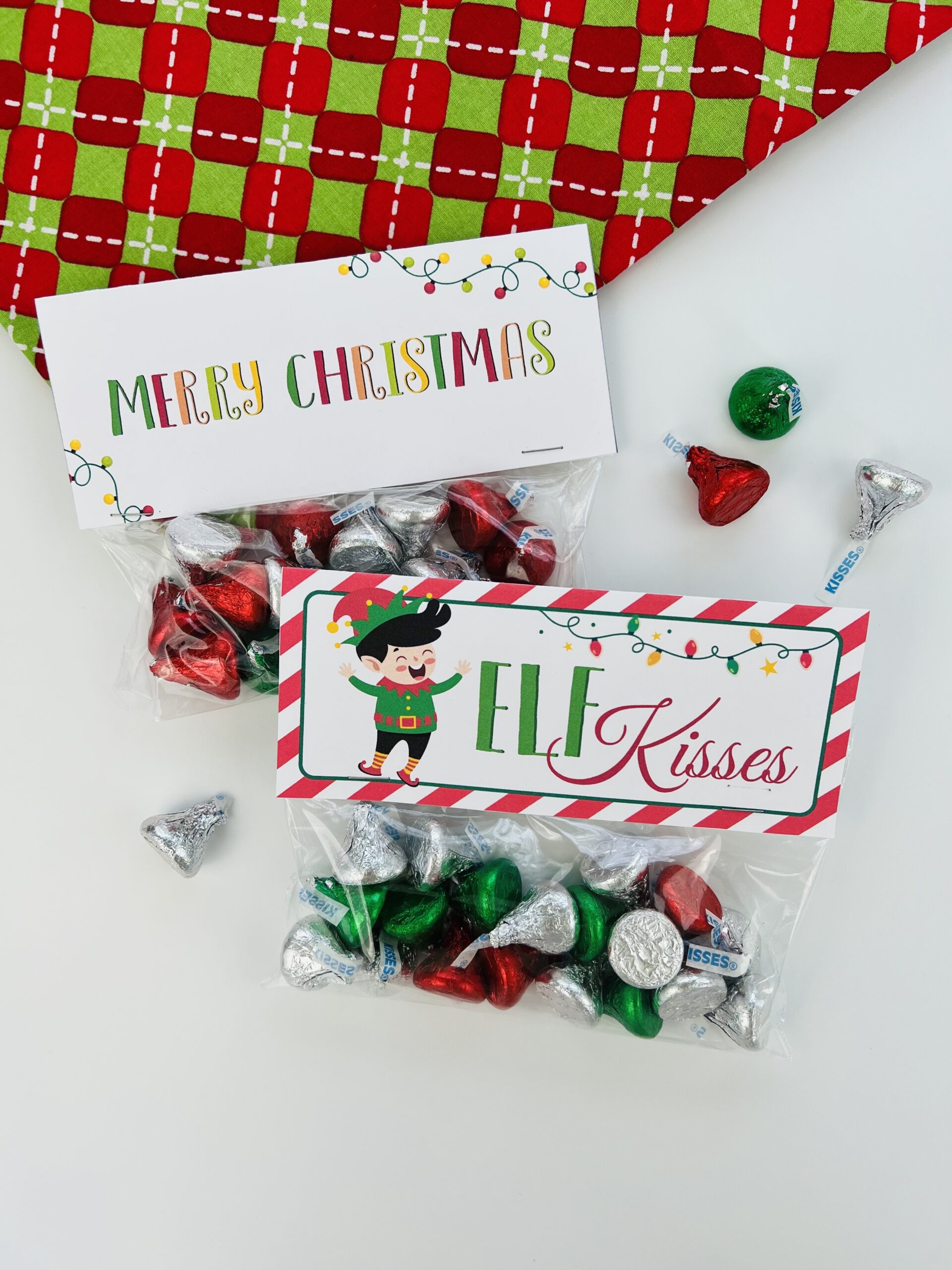 On a bright red and green background are treat bags filled with Hershey kisses. These Elf Kisses bag toppers and Merry Christmas bag toppers make the best gifts!
