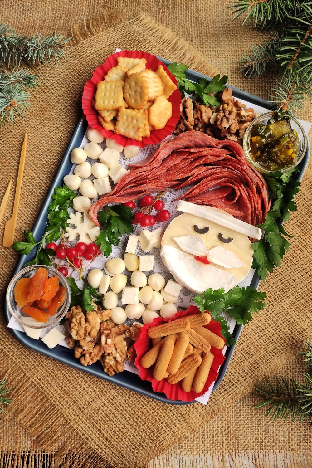 Santa Charcuterie Board. Santa is made out of brie cheese with a salami Santa hat. The Santa cheese is surrounded by other cheeses, crackers, dried fruit and nuts on a charcuterie tray.
