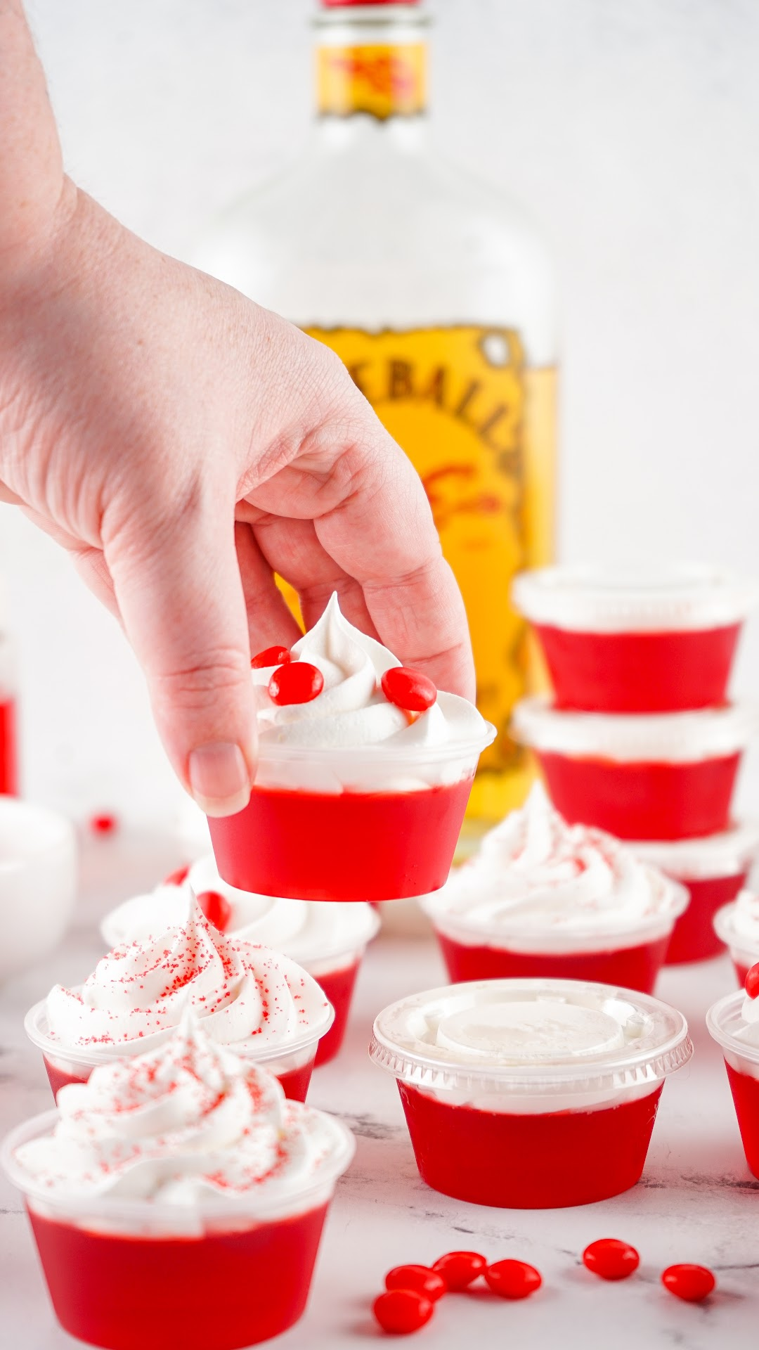 Cherry fireball jello shots with a bottle of Fireball in the background. Topped with whipped cream and red hot candy.