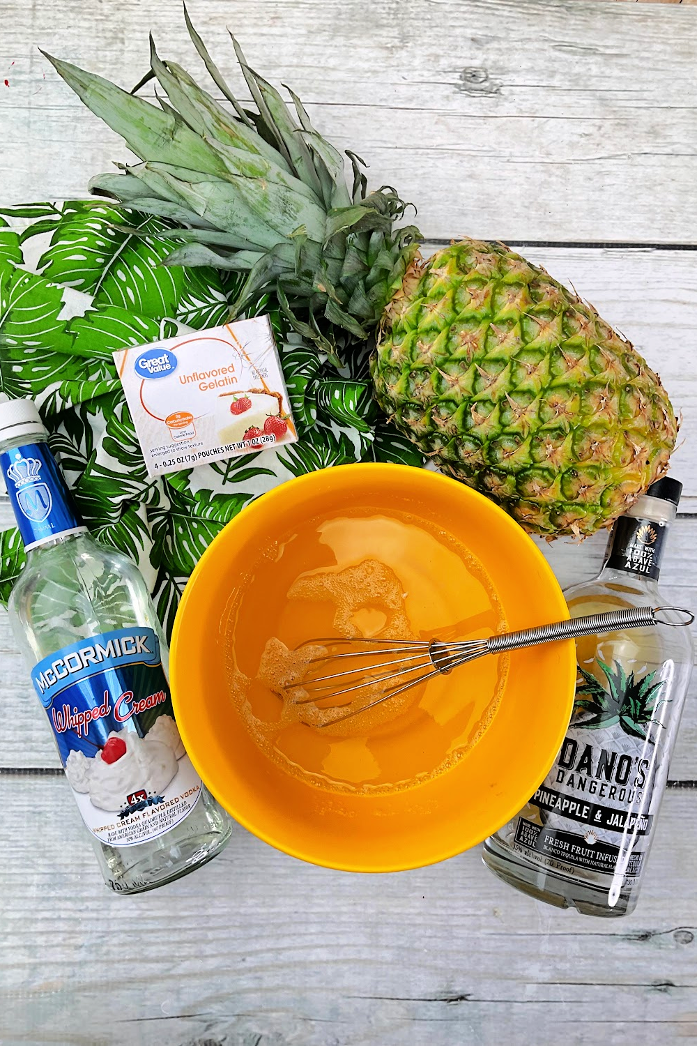 Dole whip jello shots without whip cream surrounded by ingredients. A whisk in a bowl of jello, bottles of alcohol and a pineapple.
