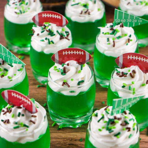 Green jello shots topped with whipped cream with brown and green sprinkles and football toppers sitting on a wood background. The perfect game day football jello shots!