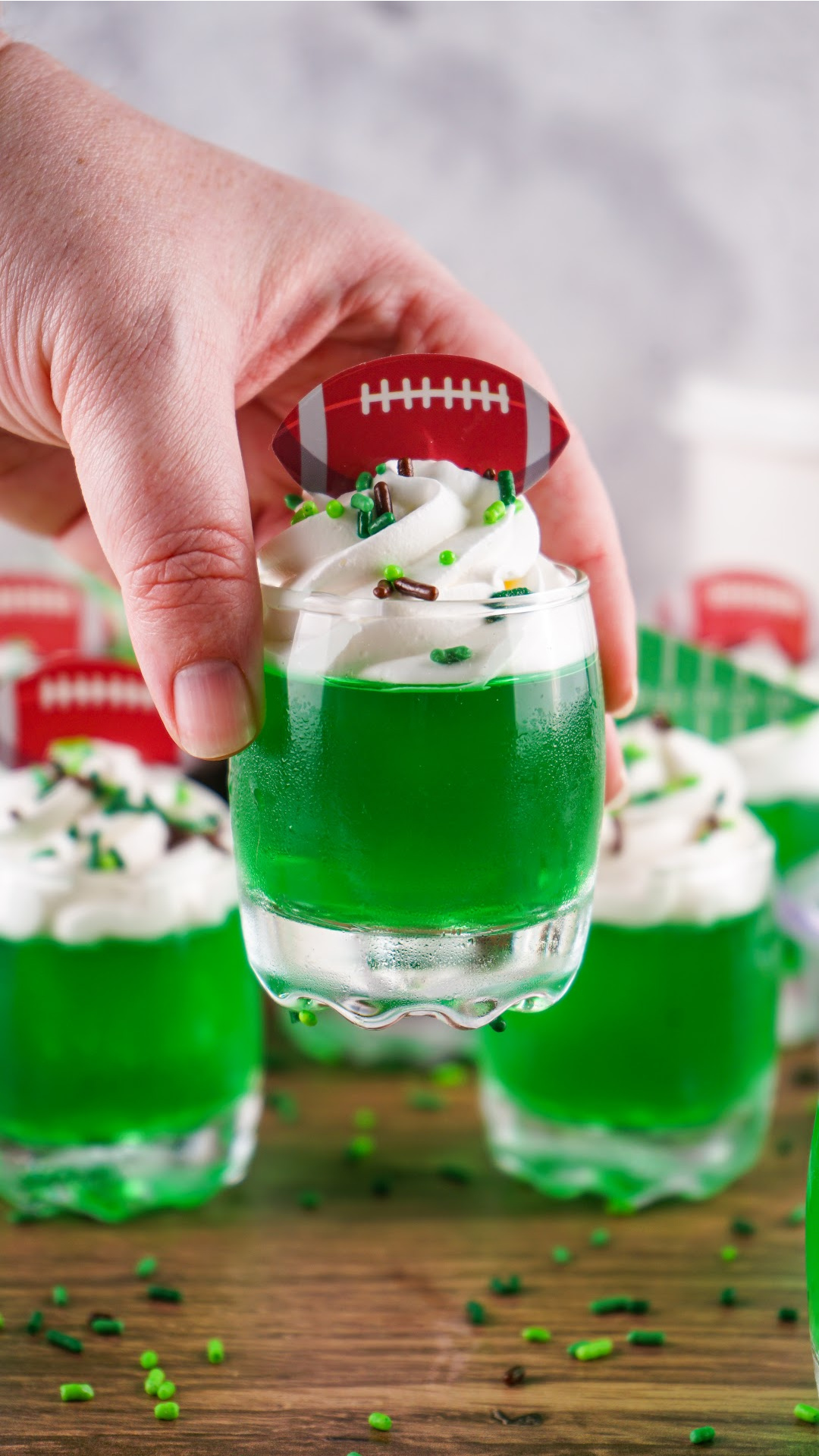 A hand holding a Green jello shots topped with whipped cream with brown and green sprinkles and football toppers. The perfect game day football jello shots!