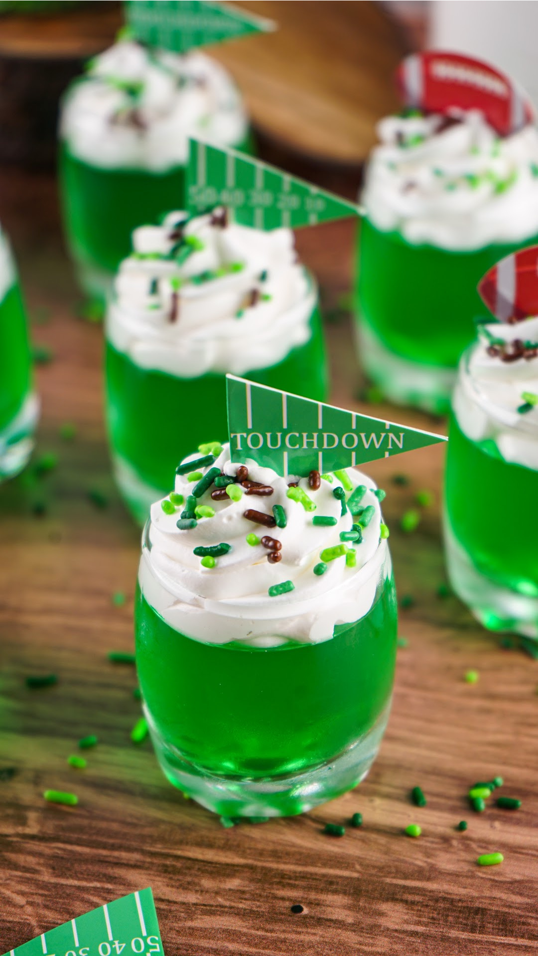Green jello shots topped with whipped cream with brown and green sprinkles and pennant toppers. The perfect game day football jello shots!