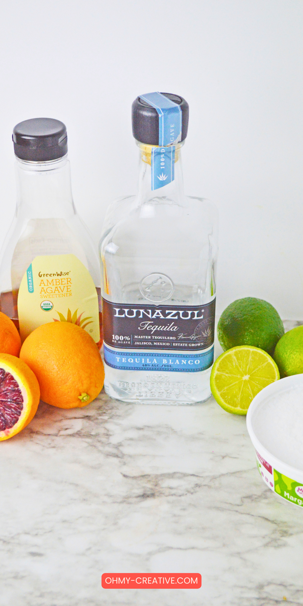 A flat lay of ingredients for a blood orange margarita on a marble surface, including a bottle of Lunazul Tequila, GreenWise organic amber agave sweetener, whole and sliced blood oranges, limes, and a container of margarita salt.