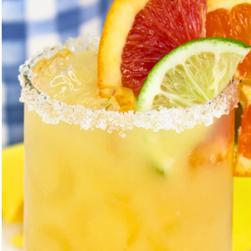 A refreshing orange margarita served in a salt-rimmed glass, garnished with slices of lime, blood orange, and orange; set on a white table with a yellow napkin, tequila bottle, and blue gingham decor in the background. Text on the image reads “Refreshing Orange Margarita"