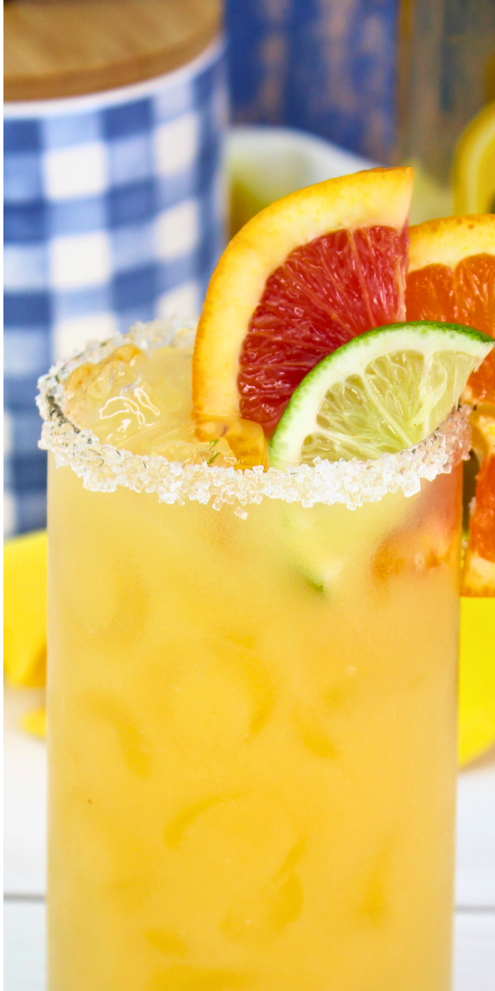 A refreshing orange margarita served in a salt-rimmed glass, garnished with slices of lime, blood orange, and orange; set on a white table with a yellow napkin, tequila bottle, and blue gingham decor in the background. Text on the image reads “Refreshing Orange Margarita"