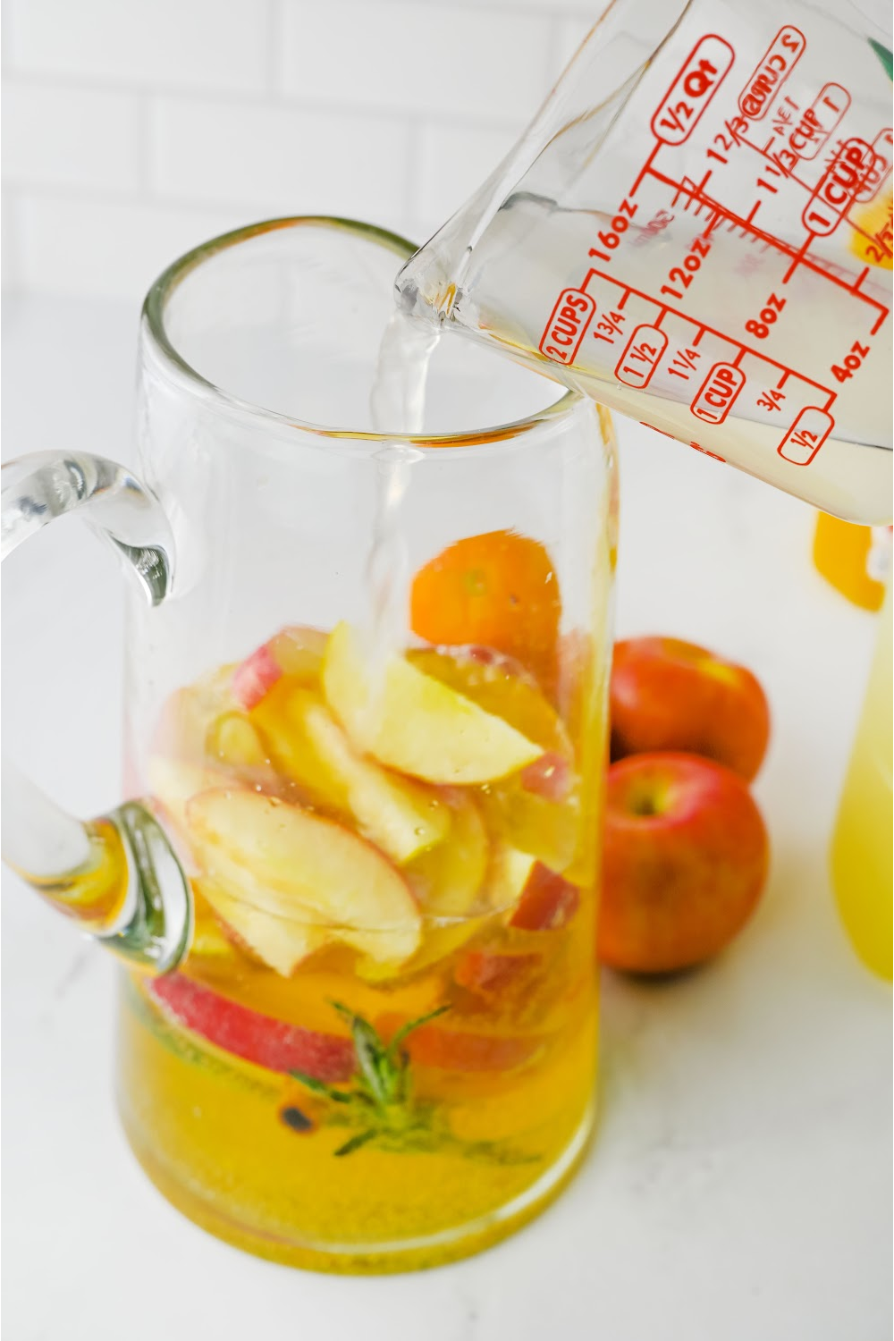 A glass pitcher with sliced fruit and a measuring cup pouring in apple cider.
