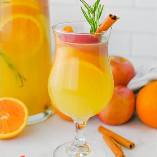 A full glass of fall punch with the pitcher of punch in the background with oranges and apples.