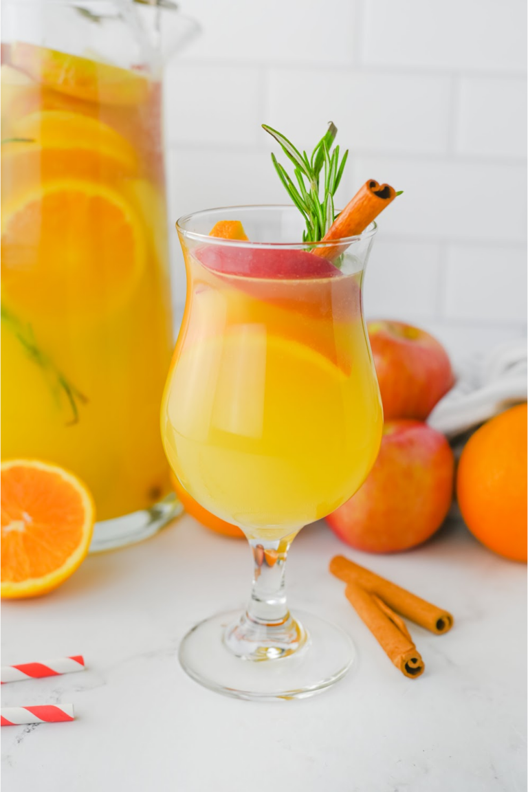 A full glass of fall punch with the pitcher of punch in the background with oranges and apples.